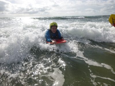 A school student on an outdoor adventure course rides a wave to the shore on her body board smiling.